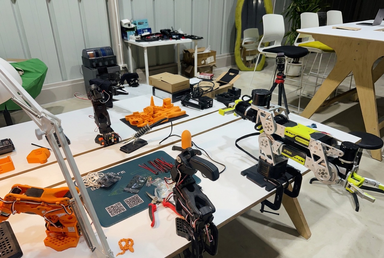 The lab bench in our studio: the reBot and the SO-101 on their clamps, orange printed parts, a camera on a tripod